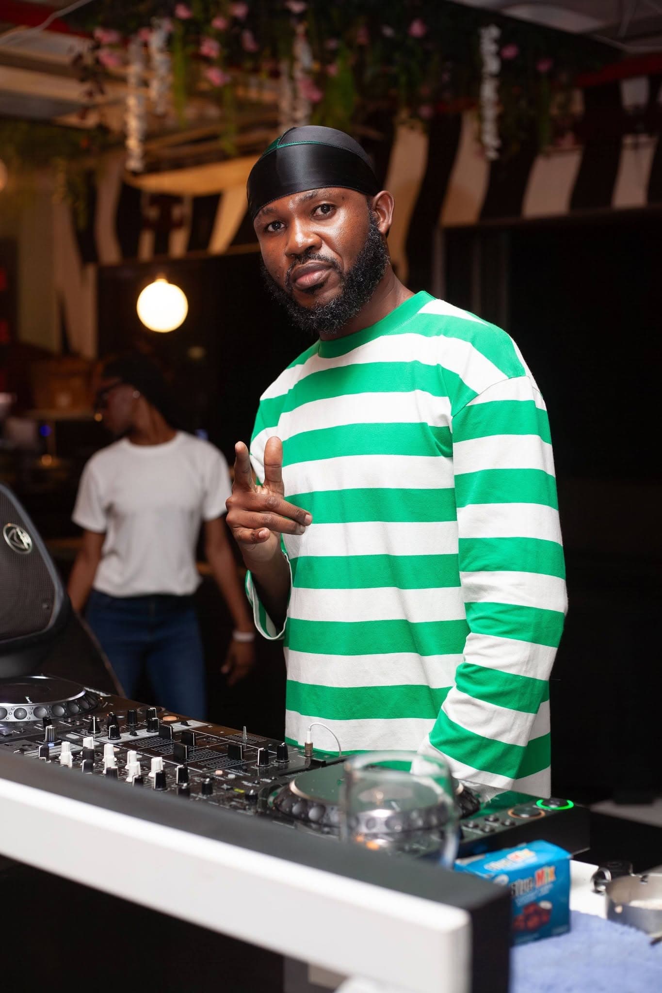 A DJ flashes a peace sign from behind the decks under warm hanging lights.
