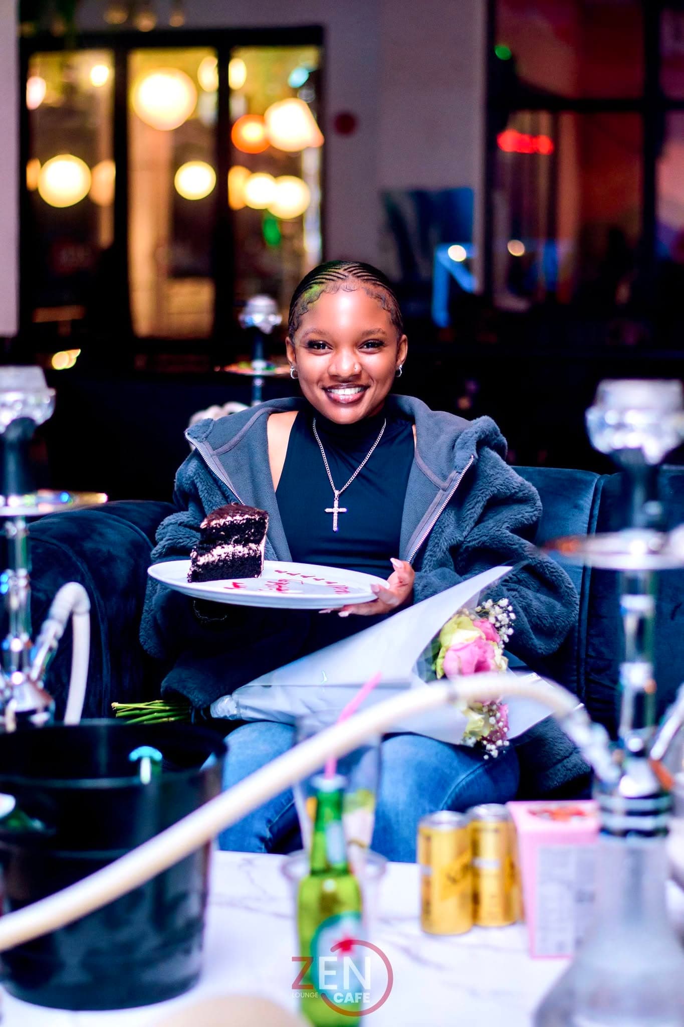 A guest smiles while holding a slice of celebration cake, hookah pipes and warm city lights behind her.