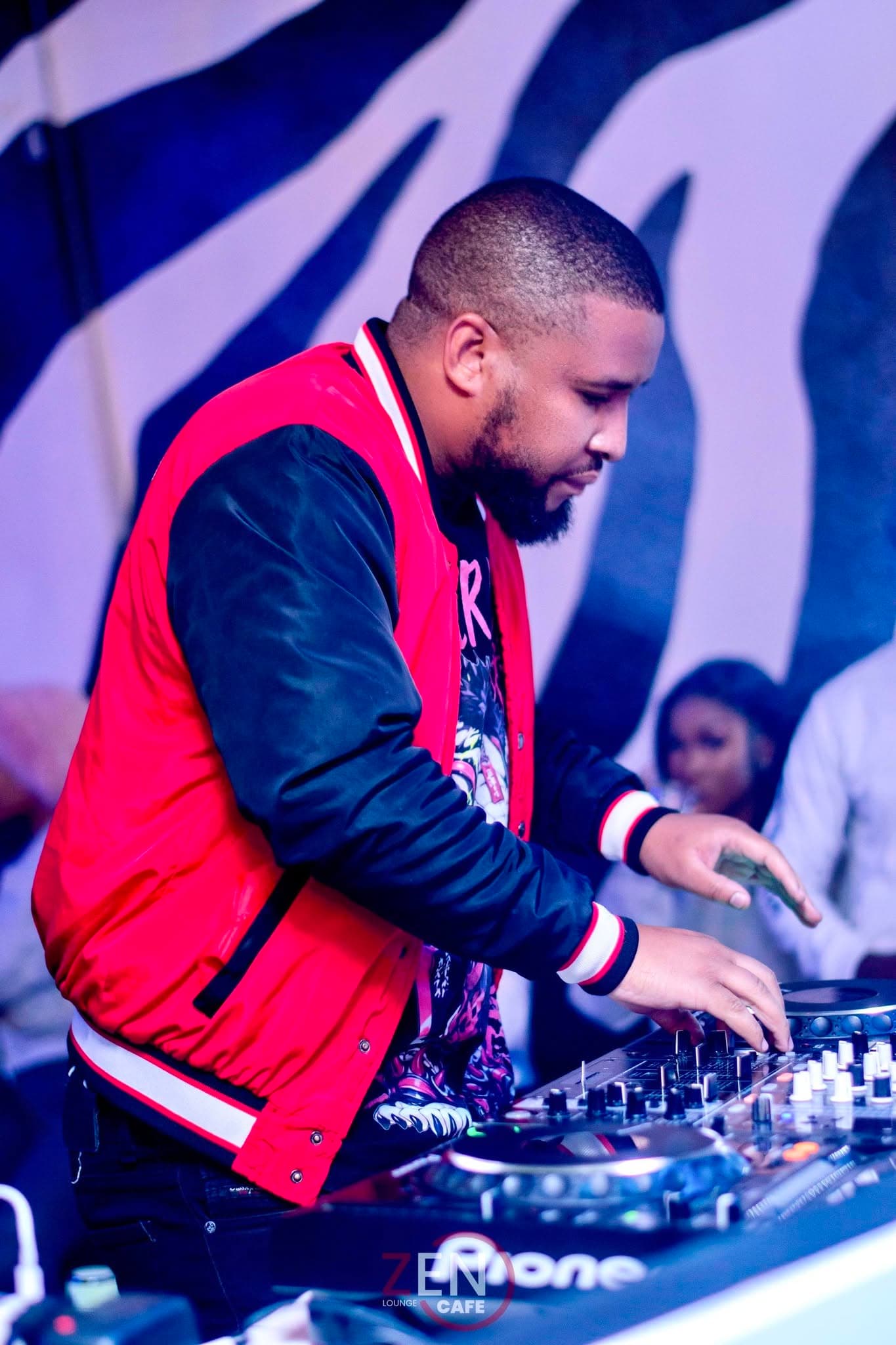 A DJ in a red racing jacket mixes tracks bathed in blue light.
