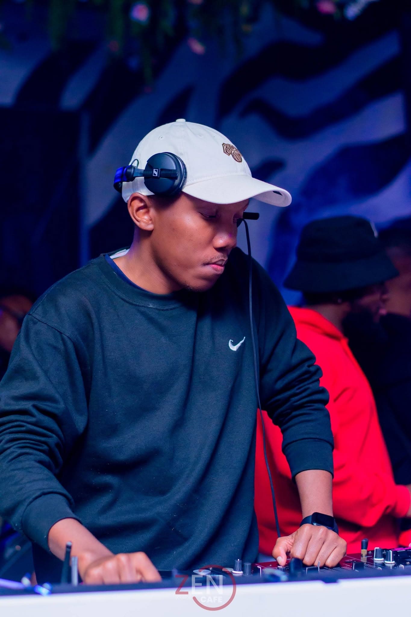A DJ focuses on the mix, headphones resting over a cap, guests dancing behind.