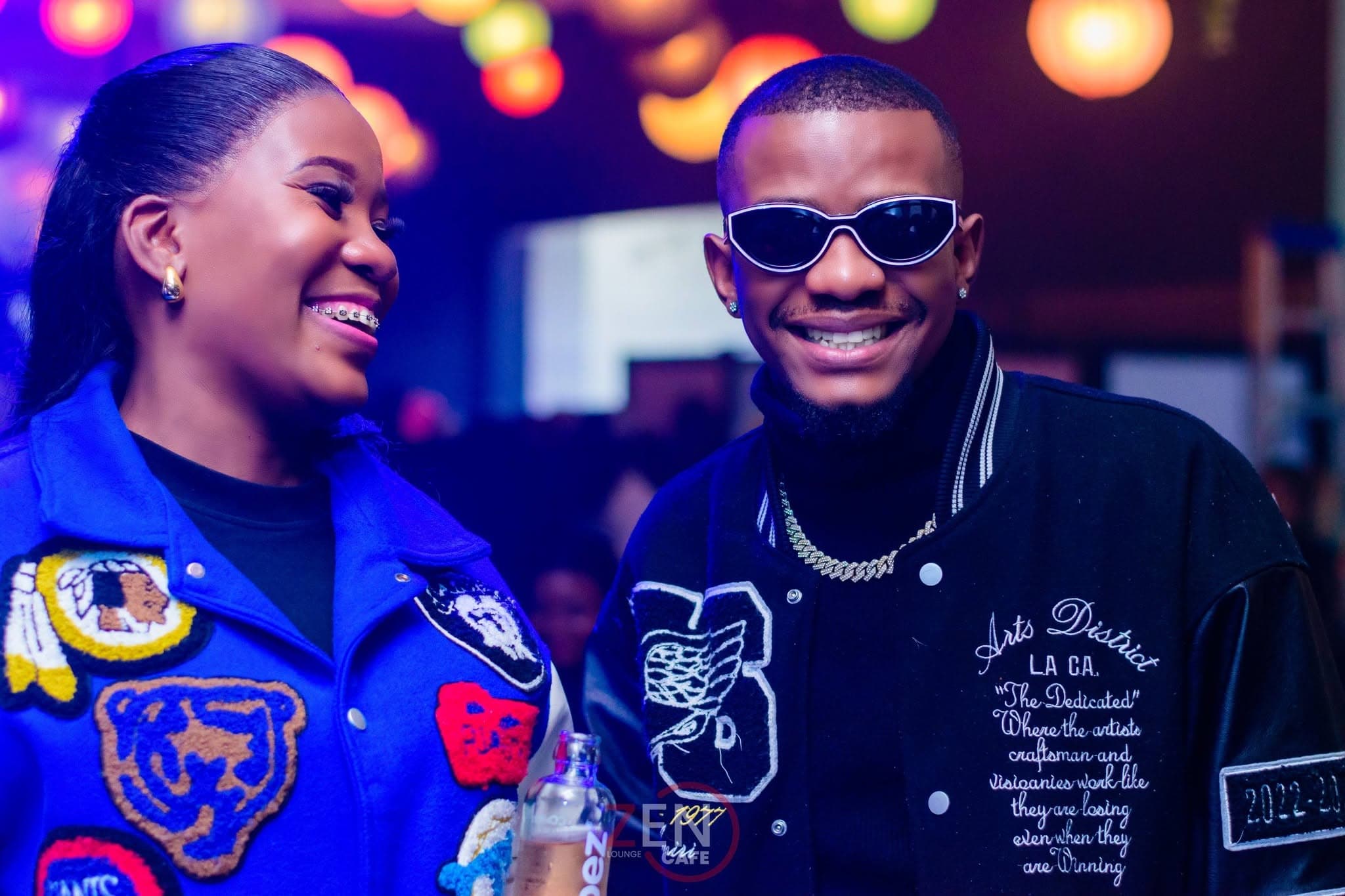 A couple share a smiling moment together, colorful lounge lights glowing behind them.