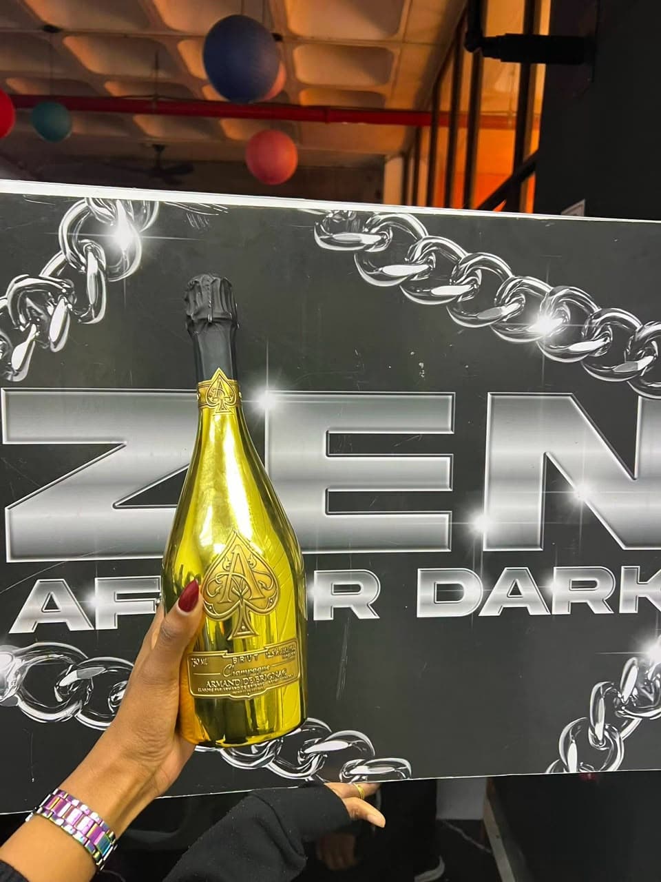 A guest holds up a gold bottle of champagne in front of the "ZEN After Dark" wall.