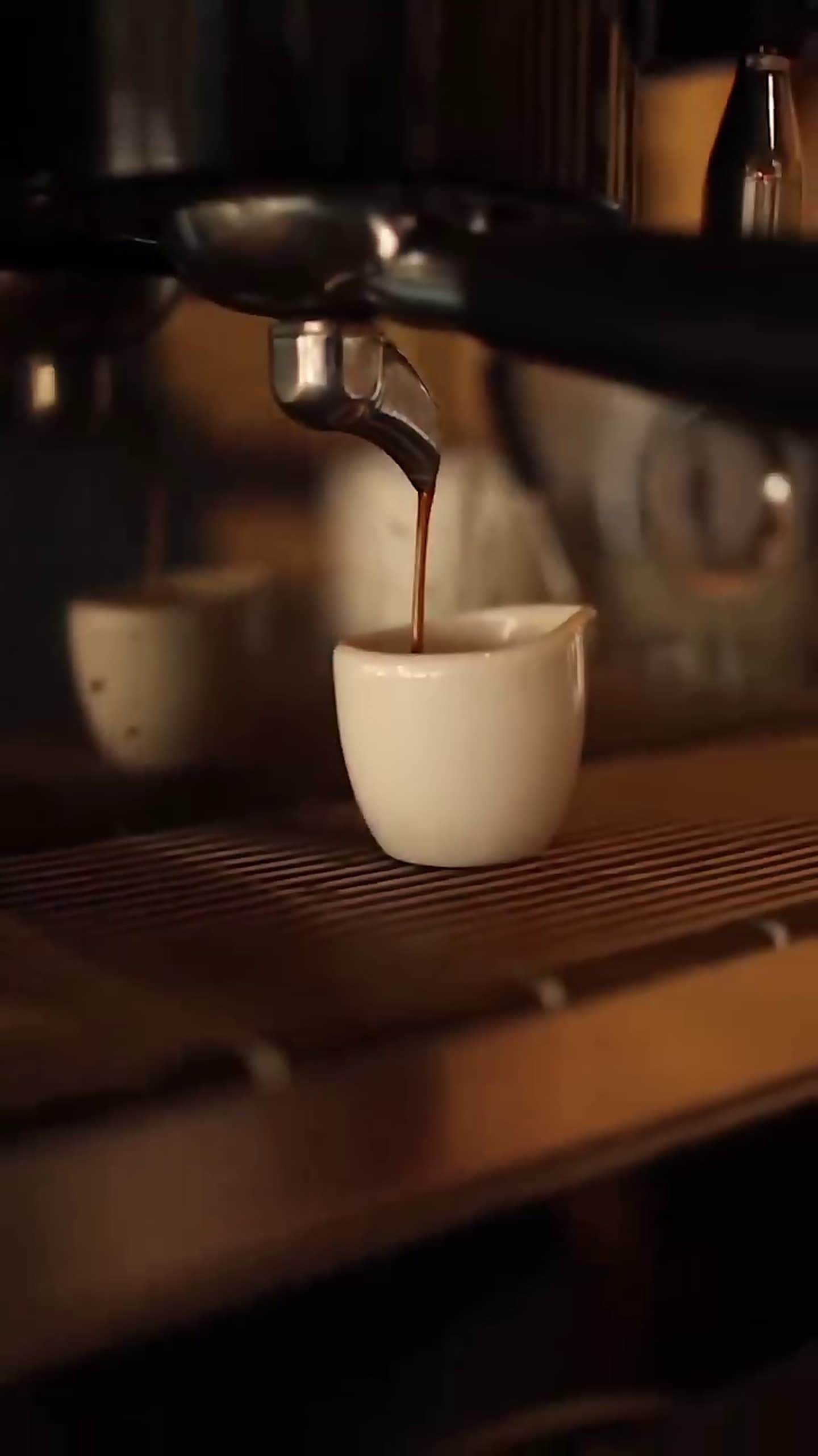 A rich espresso shot streams into a white cup beneath the group head of a professional espresso machine, warm amber light glowing behind it.