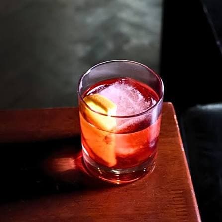 A Dusk Negroni in a rocks glass with a large ice cube and orange peel, lit in warm red light.